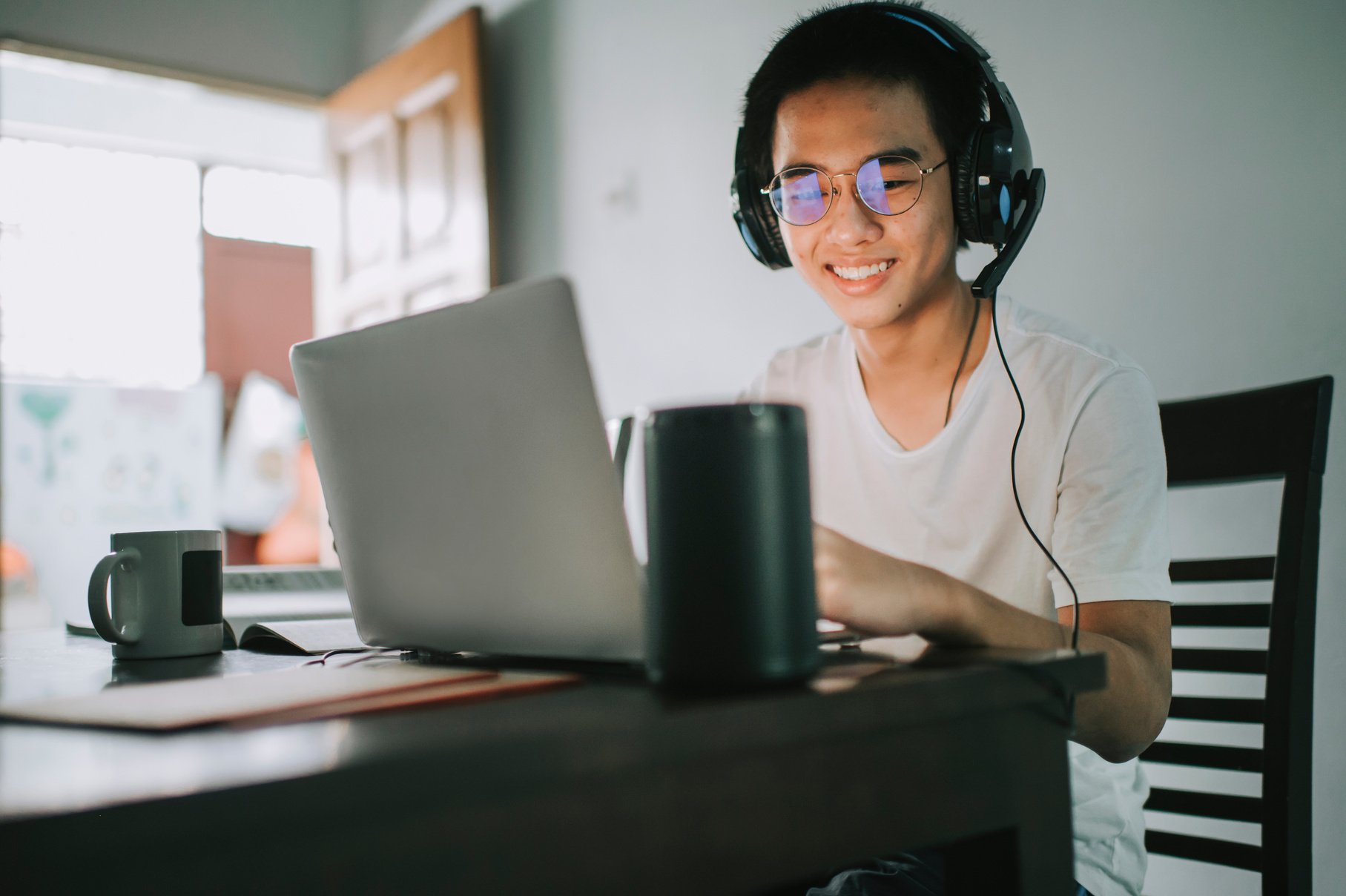 An asian chinese teenager chatting online with all his classmates via online software while doing his homework at the kitchen dining table together with his virtual assistant smart speaker